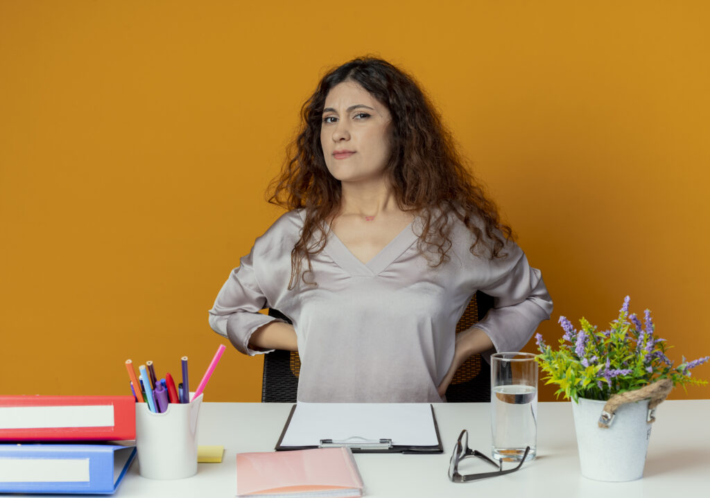 The current image has no alternative text. The file name is: tired-young-pretty-female-office-worker-sitting-desk-with-office-tools-putting-hands-back-isolated-orange-scaled.jpg